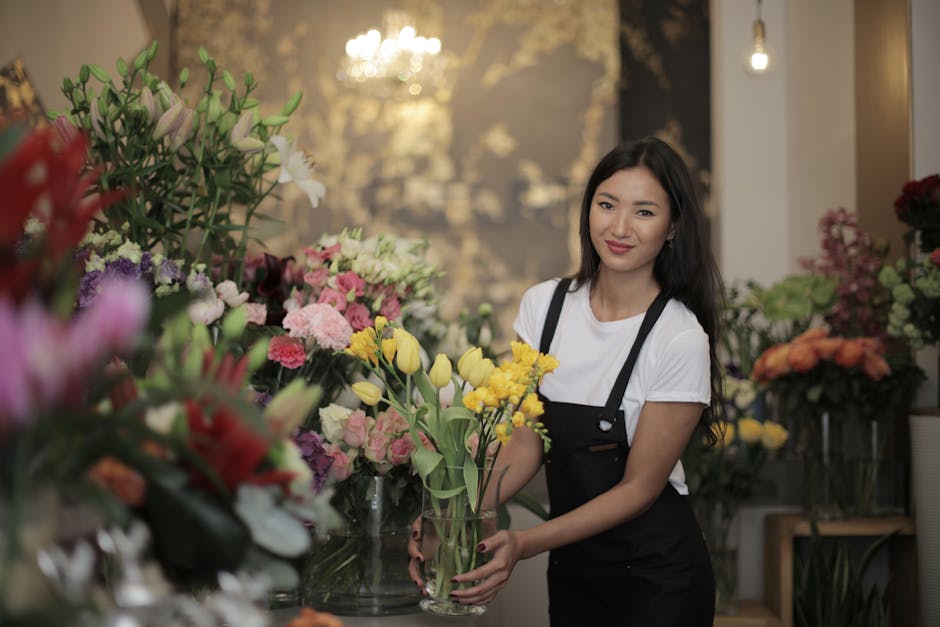 A young woman with long dark hair, wearing a white t-shirt and a black apron, is inside a florist shop, holding a glass vase filled with yellow tulips and some light pink and white flowers. The floral