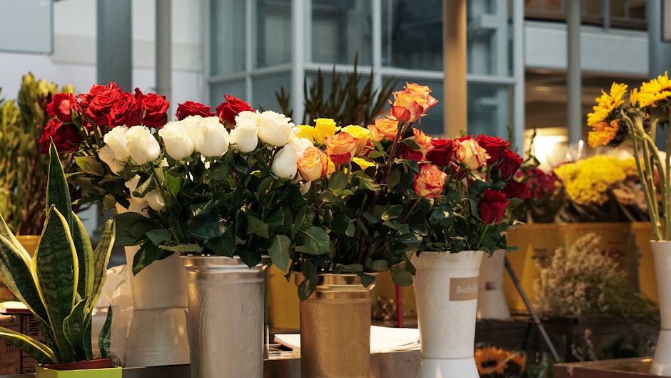 A floral display featuring three bouquets of roses arranged in metallic and white ceramic vases on a table at a flower shop. The bouquets include red, white, cream, and multicolored roses with lush gr