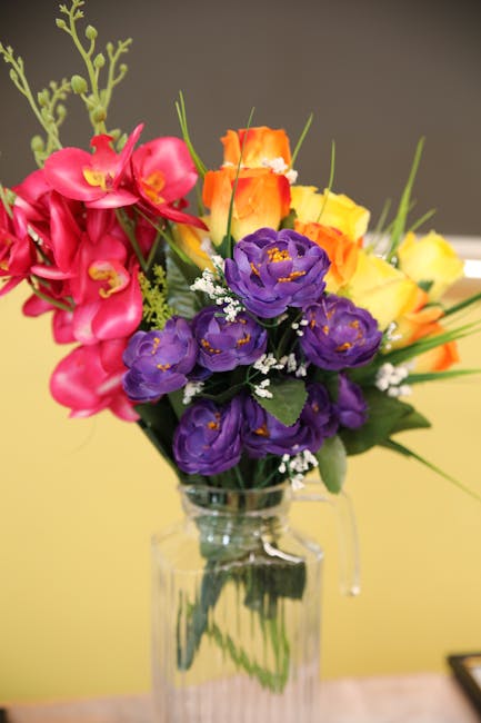 A vibrant floral arrangement featuring pink orchids with delicate petals and yellow centers, orange and yellow roses with tightly budded blooms, and deep purple eustoma flowers with ruffled petals, al