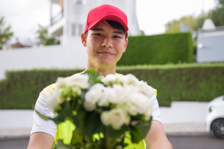 A young male flower delivery courier wearing a red cap and a fluorescent yellow vest presents a bouquet of fresh white flowers, including roses and lilies, wrapped in green foliage. The flowers are ar