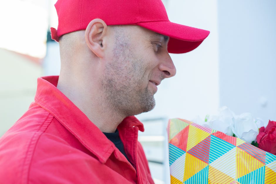 A man in a red cap and red jacket is holding a bouquet of fresh flowers wrapped in vibrant, geometric-patterned wrapping paper. The bouquet includes white blooms, possibly roses or peonies, and deep p
