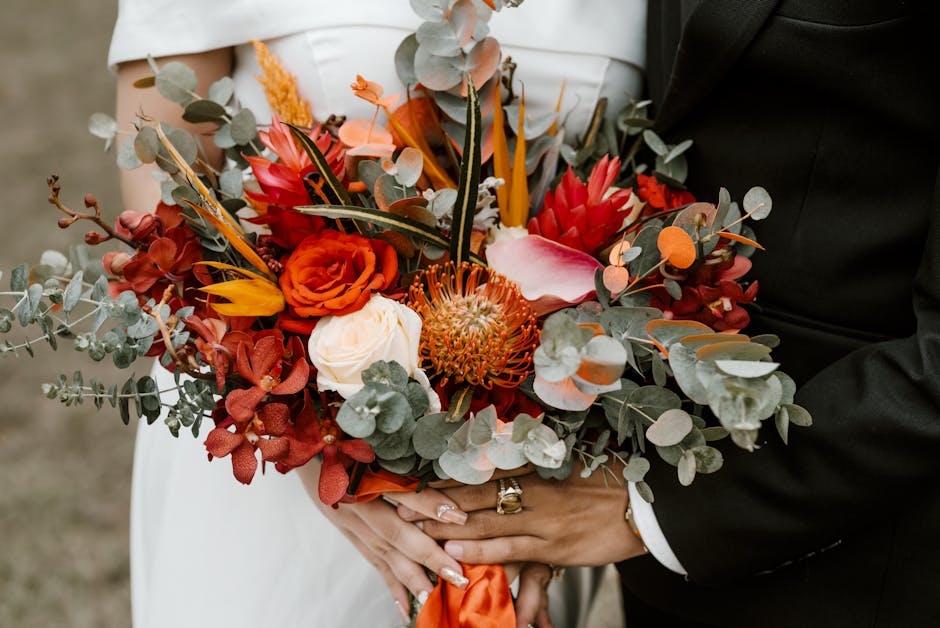 A close-up of a bridal bouquet held by a bride and groom, featuring a vibrant arrangement of flowers including orange roses, red protea, pink calla lily, and white garden roses, complemented by silver
