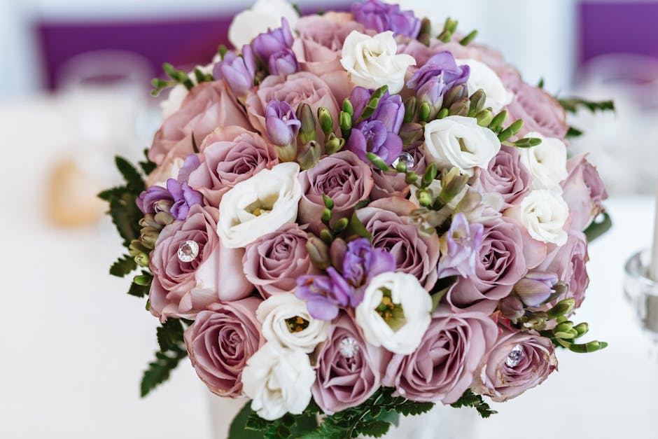 A close-up of a floral bouquet arrangement featuring soft pink roses, white lisianthus, and purple eustoma, accented with small green buds and foliage. The bouquet is beautifully crafted with a lush,
